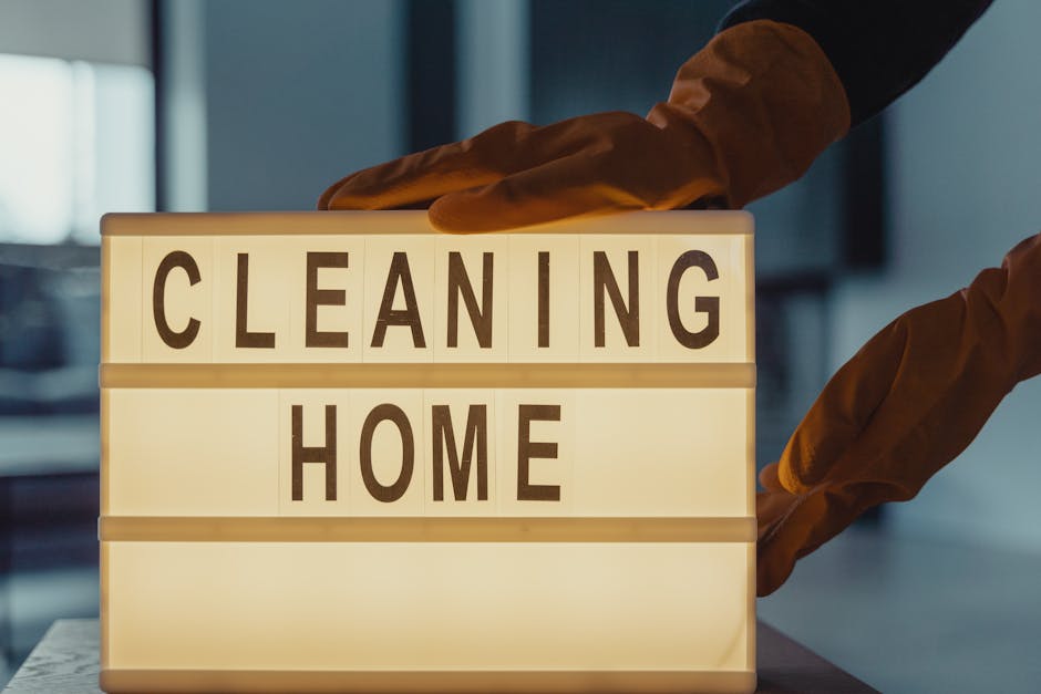 A close-up of a lightbox on a wooden table displaying the words 'CLEANING HOME' in black letters. The background shows a modern, well-lit interior with dark-colored walls, a large window allowing natu
